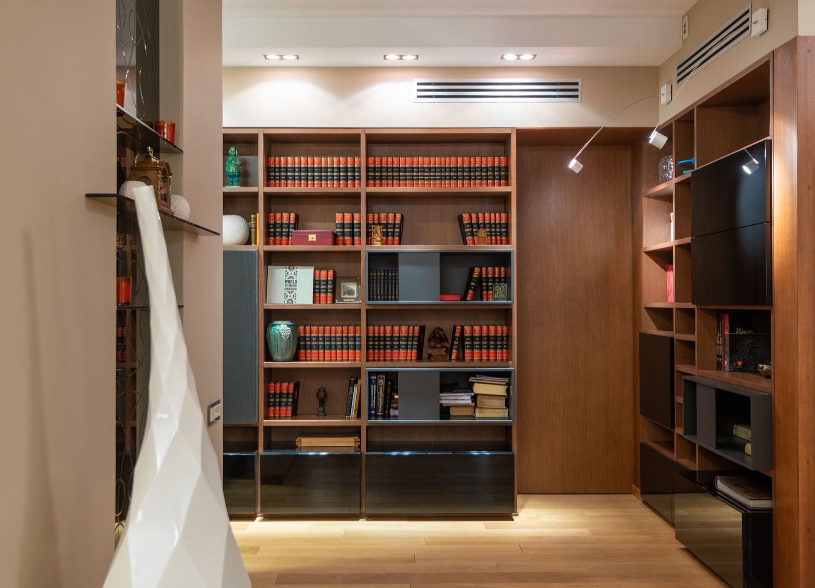 Built-in walnut bookcase wall with rows of matching hardbound volumes, a few ceramic pieces, grey open boxes and black glass-front cabinets at the base, lit by ceiling downlights and small adjustable wall-mounted reading lamps
