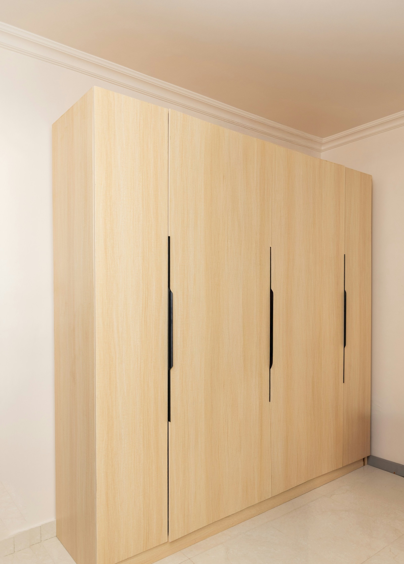 Four-door floor-to-ceiling wardrobe in a pale oak wood-grain laminate finish with long black recessed pull handles, under a white cornice on a cream marble floor