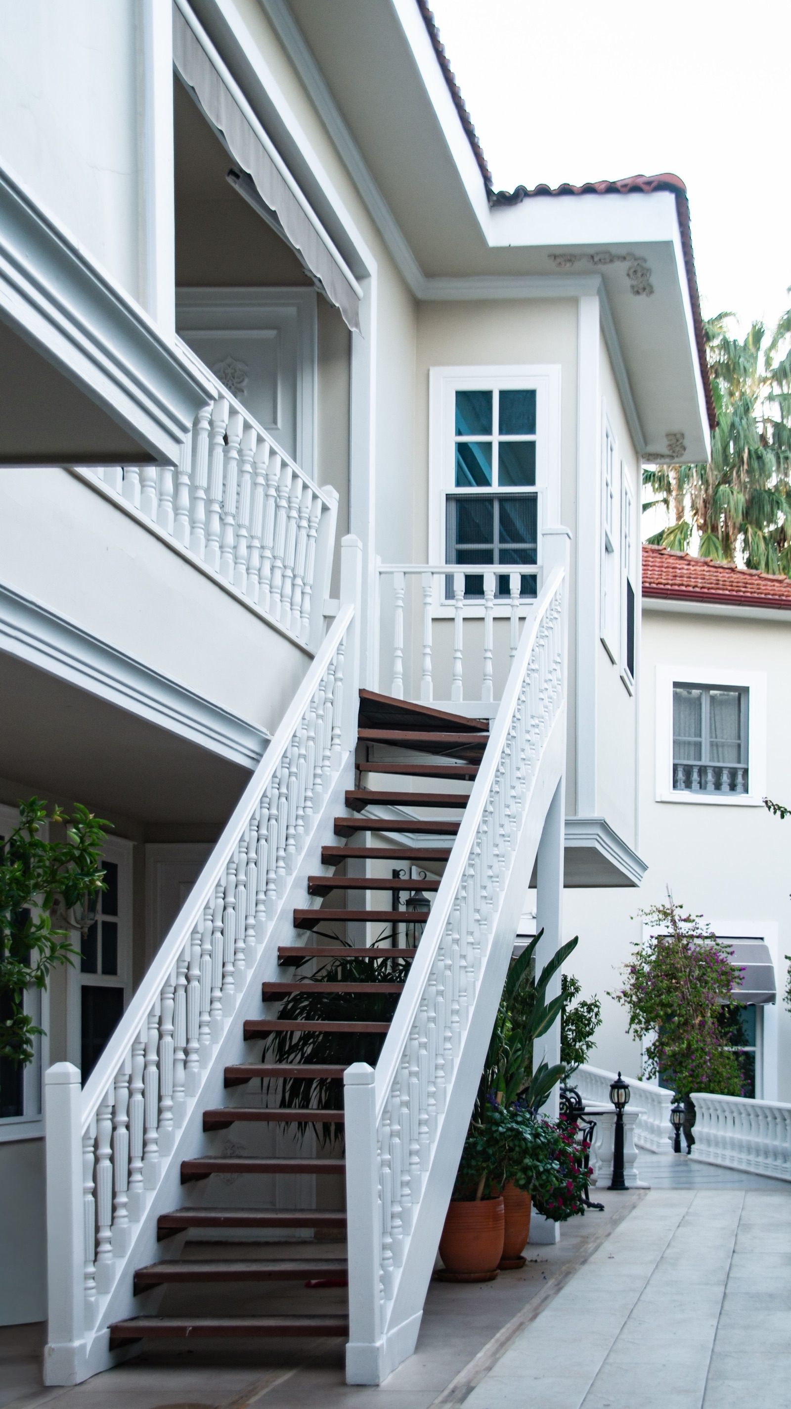A straight external staircase with dark timber treads and open risers rising along a white two-storey villa to an upper verandah, with white turned balusters on both sides and potted plants beneath the flight