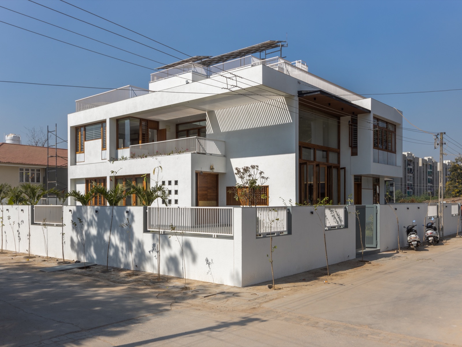 Photograph of a two-storey white bungalow on a corner plot in Ahmedabad, with textured white plaster walls, timber-framed windows and doors, a deep shaded balcony under white louvres, solar panels on a frame over the roof terrace and a white boundary wall along the street.
