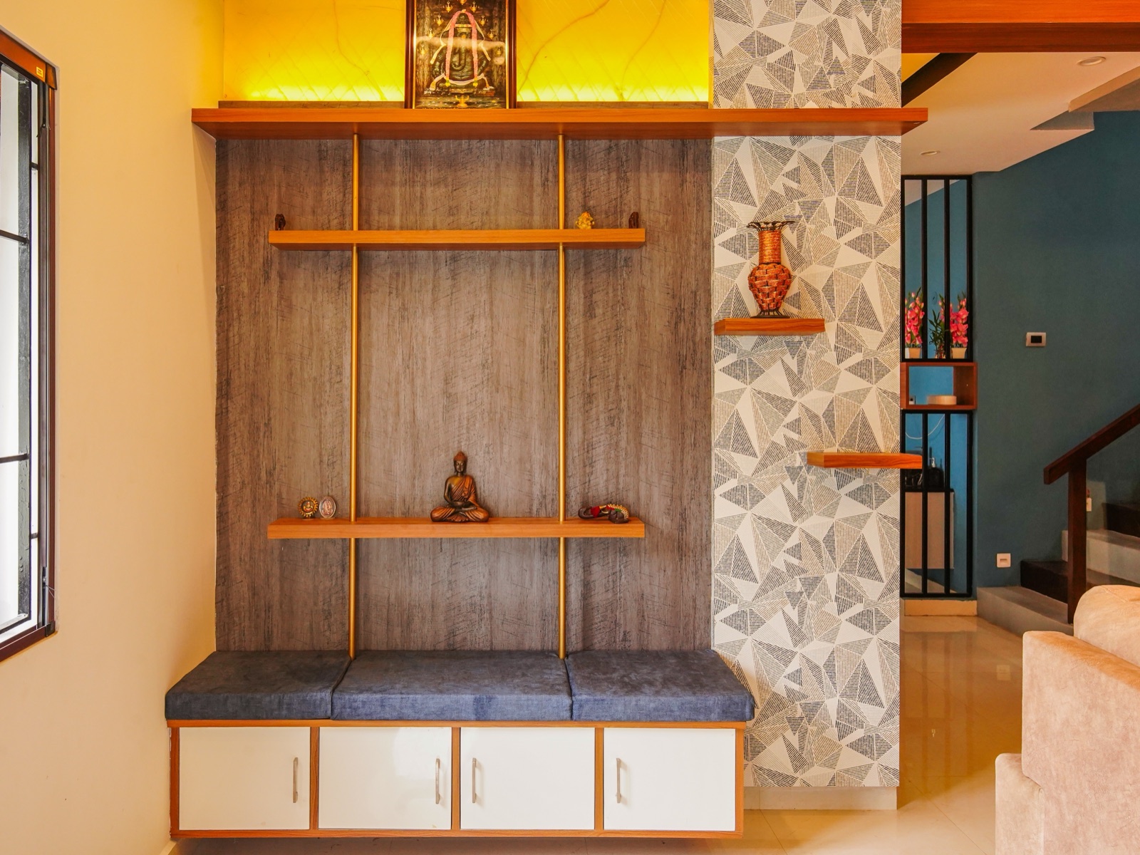 A foyer wall unit in a Chennai home: a grey wood-texture back panel with two floating wooden shelves held by slim brass rods, a small seated Buddha figure and small devotional pieces on the lower shelf, a wide top shelf lit from behind by a glowing yellow onyx-look panel that frames a garlanded devotional picture, and a long cushioned grey bench over four white cupboards at floor level. Geometric wallpaper and a staircase are to the right.