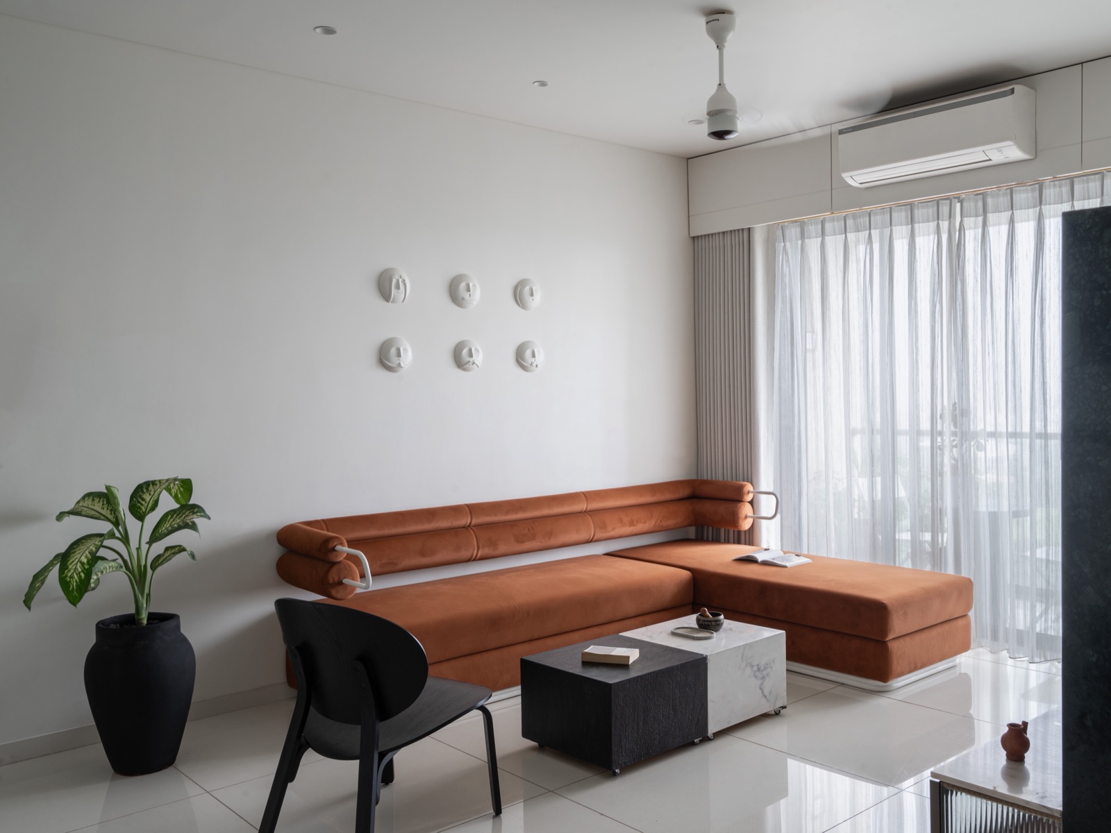 A minimal living room with white walls and a polished white tile floor, a rust-orange velvet L-shaped sofa set against a solid wall, a black and marble block coffee table, a black chair, a potted plant, and sheer curtains over a full-height window