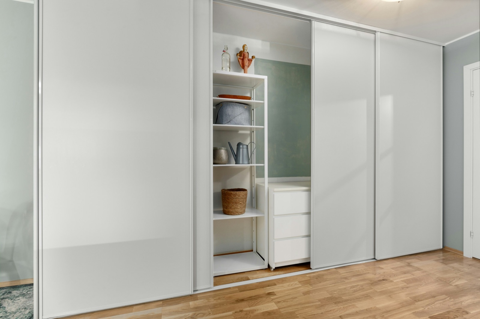 Wall-to-wall wardrobe with tall white sliding panels in slim aluminium frames, one panel pushed aside to show open white shelves and a three-drawer unit inside, on an oak floor