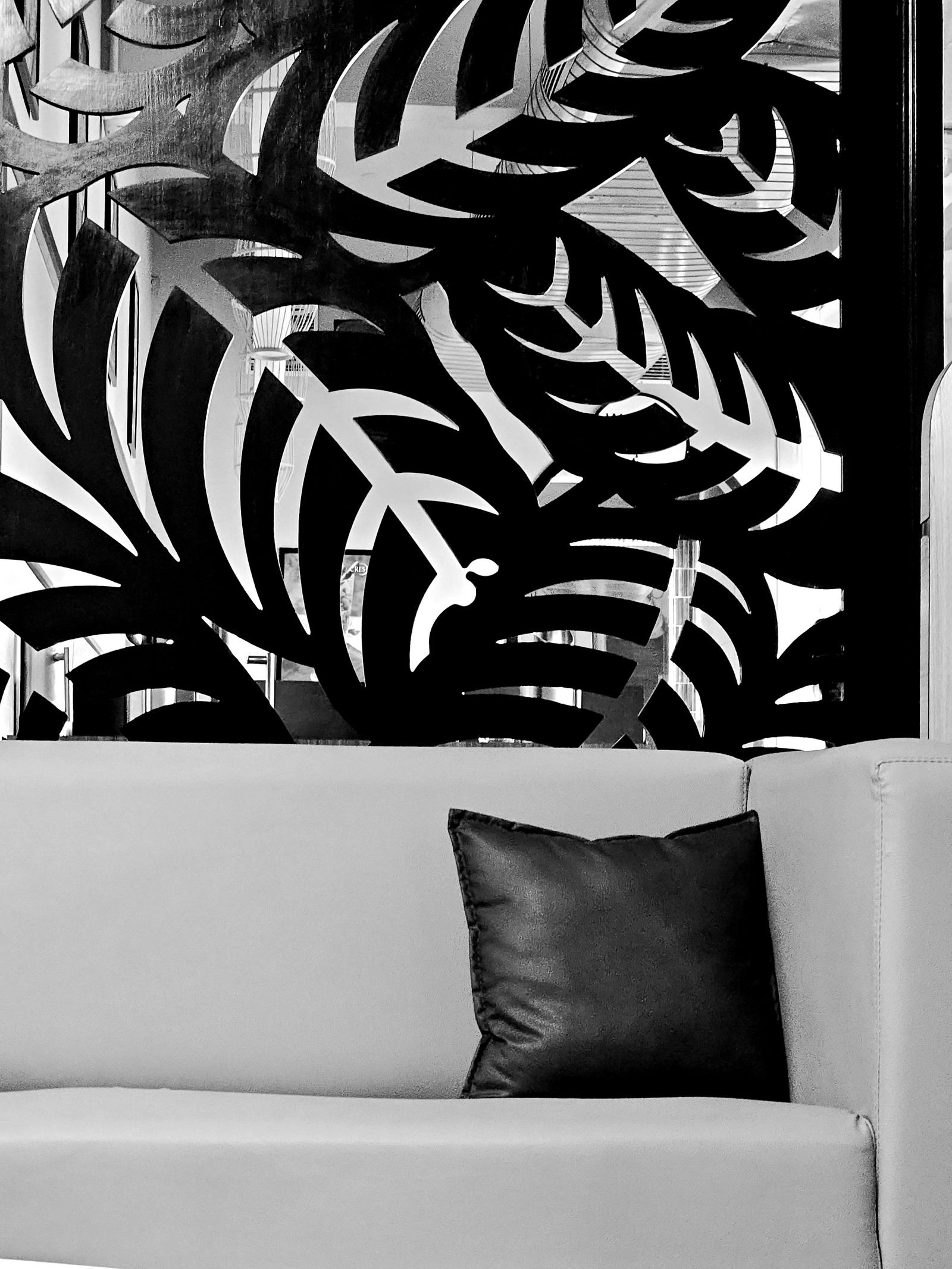 Black and white photo of a large laser-cut panel with a bold pattern of leaves and fronds standing behind a pale leather sofa with one dark cushion, with the room beyond visible through the cut-outs.
