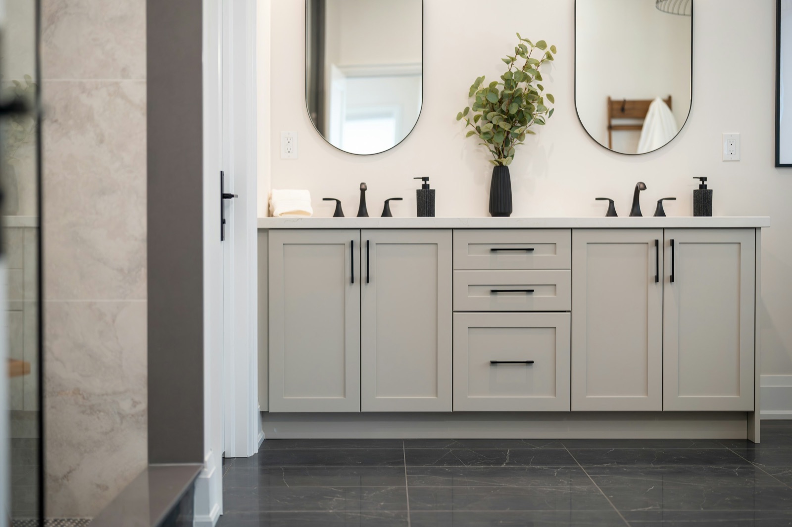 Double vanity with pale grey shaker cabinets, a white stone counter and two matt black three-hole basin mixers under oval mirrors, with a dark marble-look floor