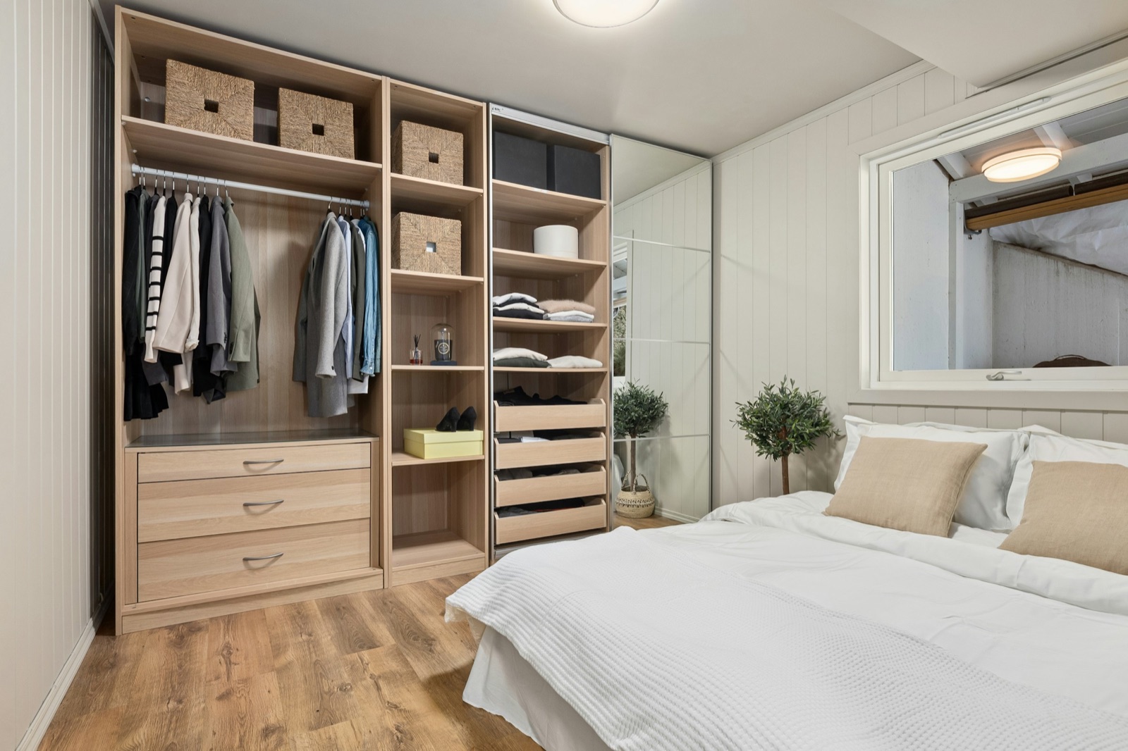 Bedroom with an open wardrobe system in light oak fitted into a recess between two walls, with hanging rails, open shelves with woven baskets, a stack of pull-out drawers and a full-height mirrored sliding door beside it, next to a bed with white linen