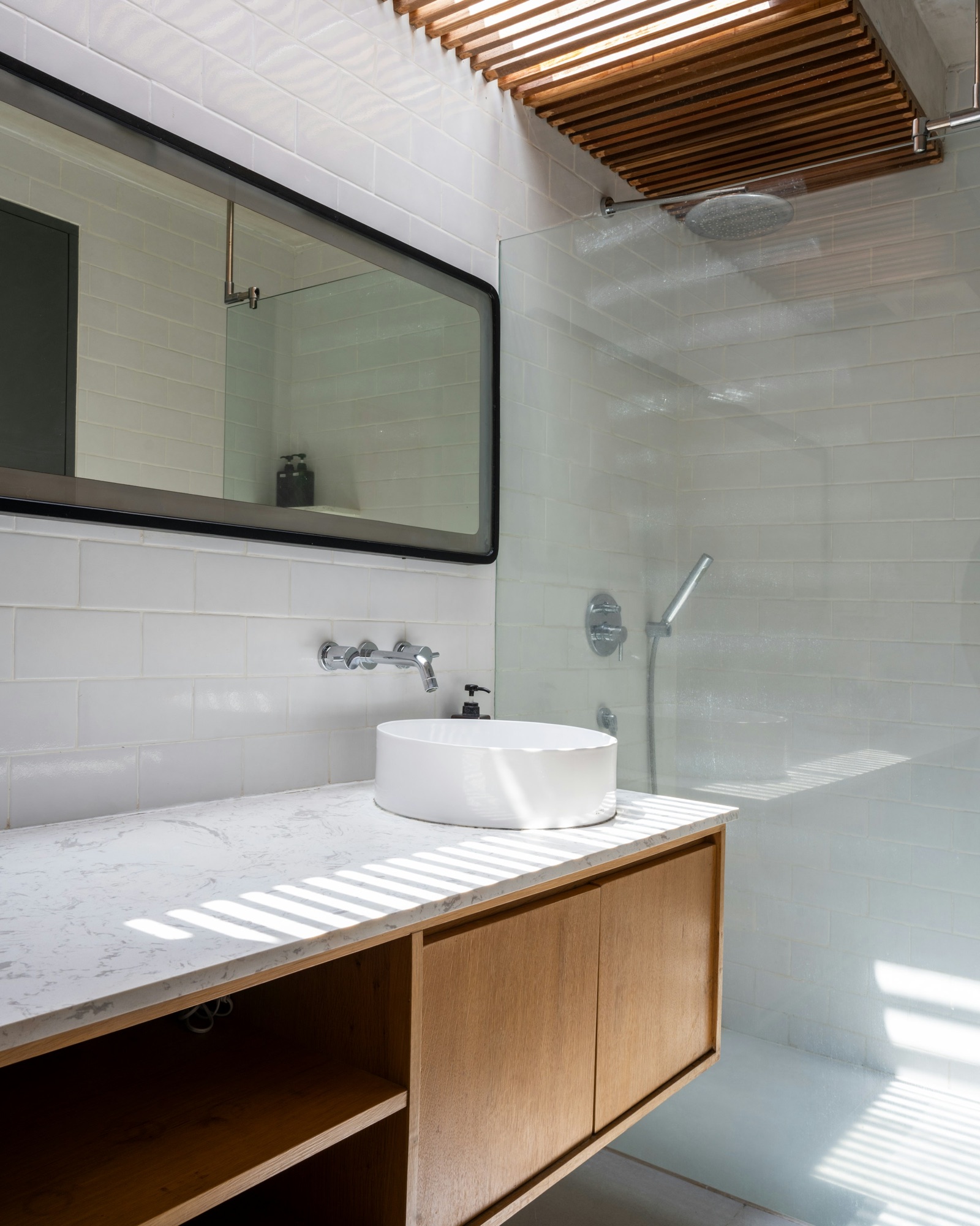 A sunlit bathroom with white brick-pattern wall tiles: a wide black-framed rectangular mirror above a floating oak vanity with a white quartz top and a round vessel basin, a wall-mounted chrome tap, and a frameless glass shower screen beside it under a timber-slatted skylight that casts stripes of sun across the counter.