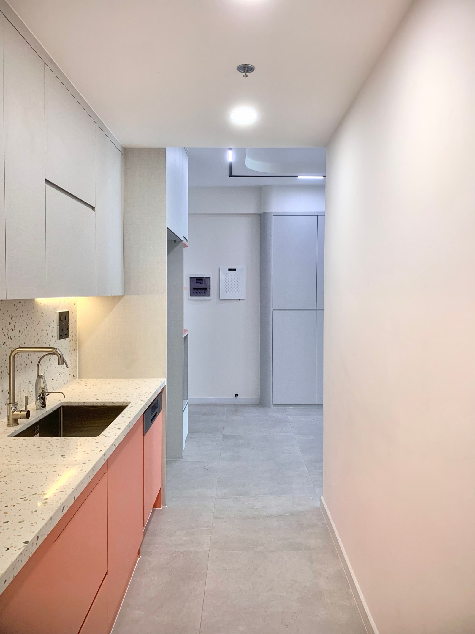 Narrow galley kitchen with coral base units, a white terrazzo-look counter and backsplash, and light grey wall cabinets. The floor is large-format grey stone-look tile with few joints, running from the kitchen into the passage beyond.