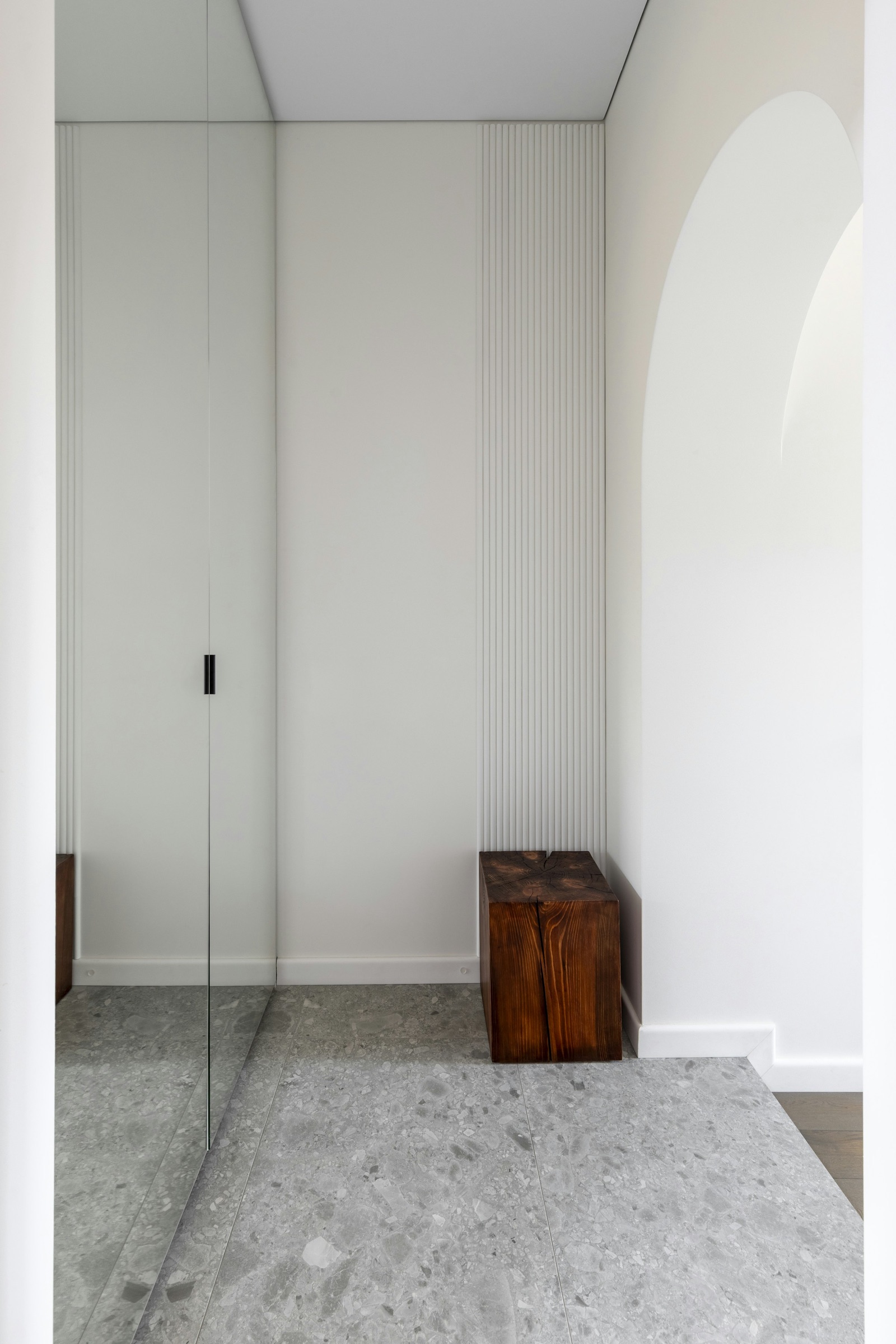 A small white entrance hall in an apartment with floor-to-ceiling mirrored doors on a built-in cupboard on the left, a fluted white wall panel, a solid wood block stool and grey terrazzo floor tiles, with an arched opening to the right; the mirror doubles the floor and wall.