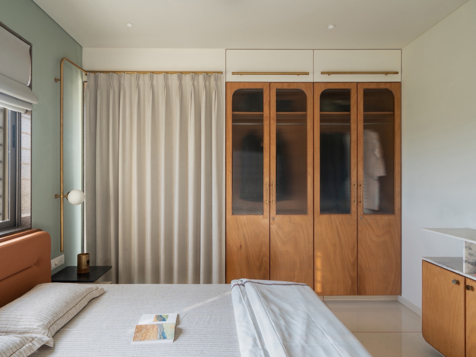 Photograph of a bedroom wardrobe in Rajkot with four wood-finish shutters whose upper halves are reeded glass, showing a hanging rail and a shelf inside, with a white two-door loft above and a brass pipe lamp beside the bed.