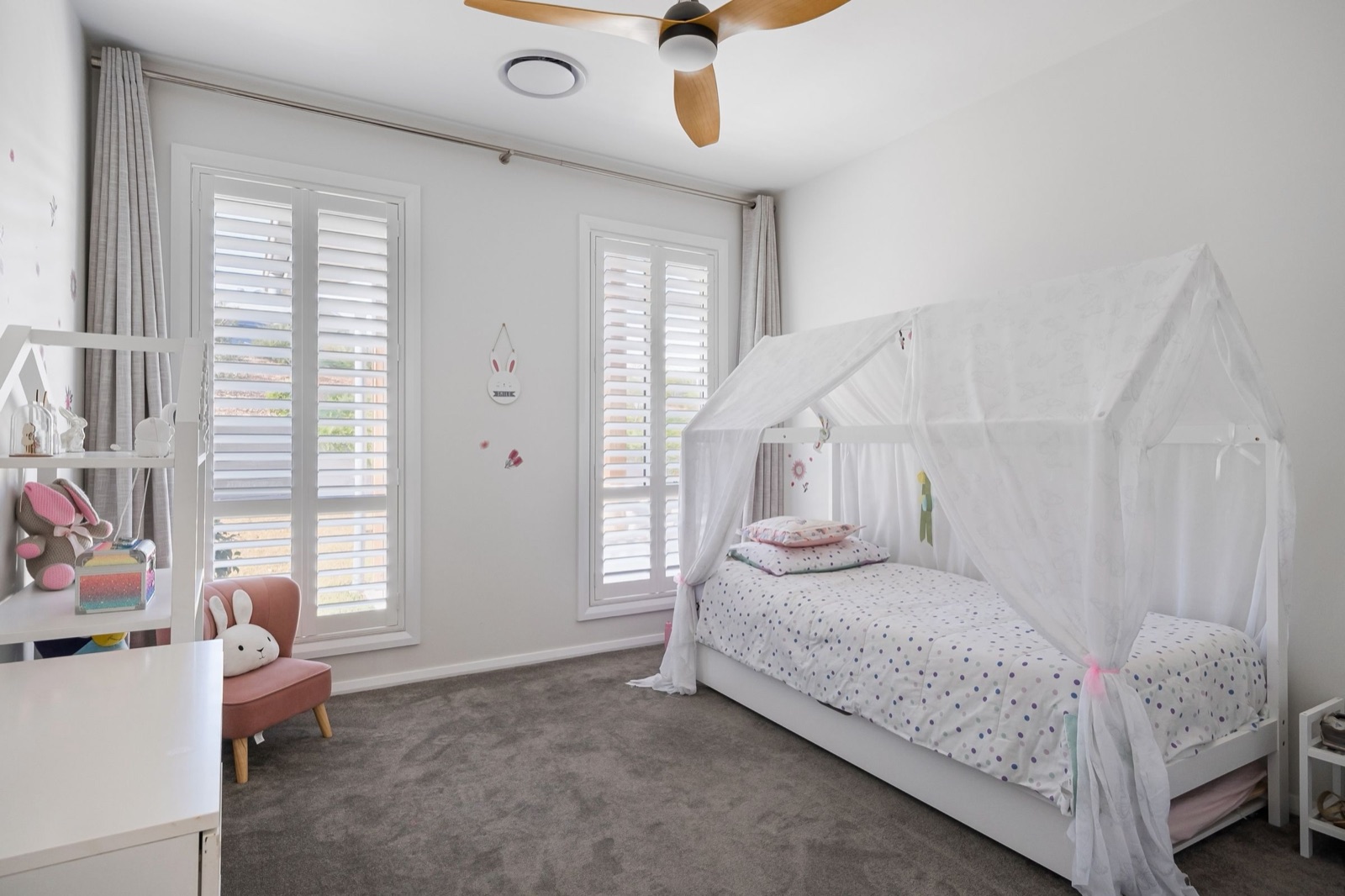 A white house-shaped single bed draped in sheer curtains, with a pull-out base below it, in a light grey kids room with plantation shutters on two tall windows, a ceiling fan, a small pink armchair and white open shelves.