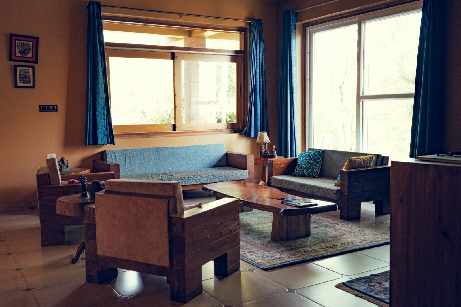 A living room with chunky solid wood sofas and armchairs with blue and grey cushions, a live-edge wooden coffee table on a patterned rug, large timber-framed windows with blue curtains and a terracotta-coloured wall