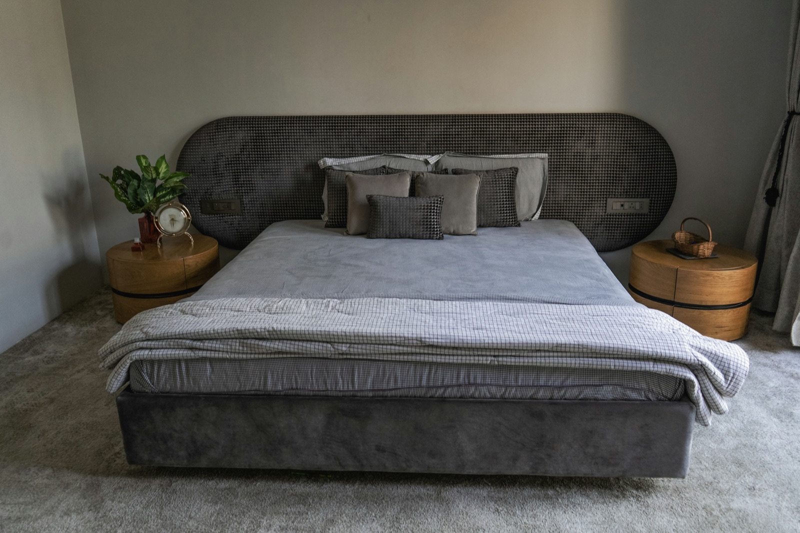 A double bed with a grey velvet base and a wide, rounded grey quilted headboard that runs past both sides of the bed, a modular switch plate set into the headboard on each side, round wooden bedside tables on both sides with a clock and plant on one and a small basket on the other, and a grey carpet.