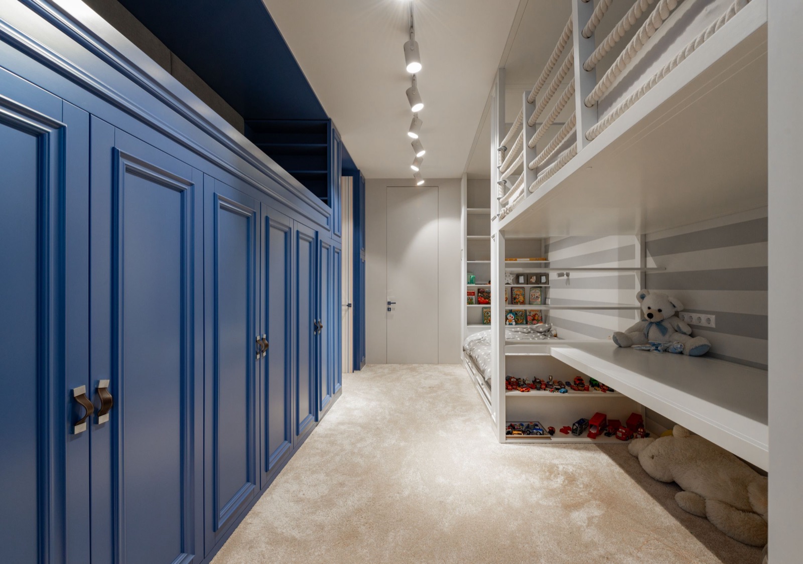 A built-in white bunk in a kids room: the upper bed is fenced with rope-wrapped guard rails, a lower bed sits behind open shelves of picture books, a long bench shelf runs under the upper bunk with low shelves of toy cars beneath, and full-height blue panelled wardrobes line the opposite wall under track lights.