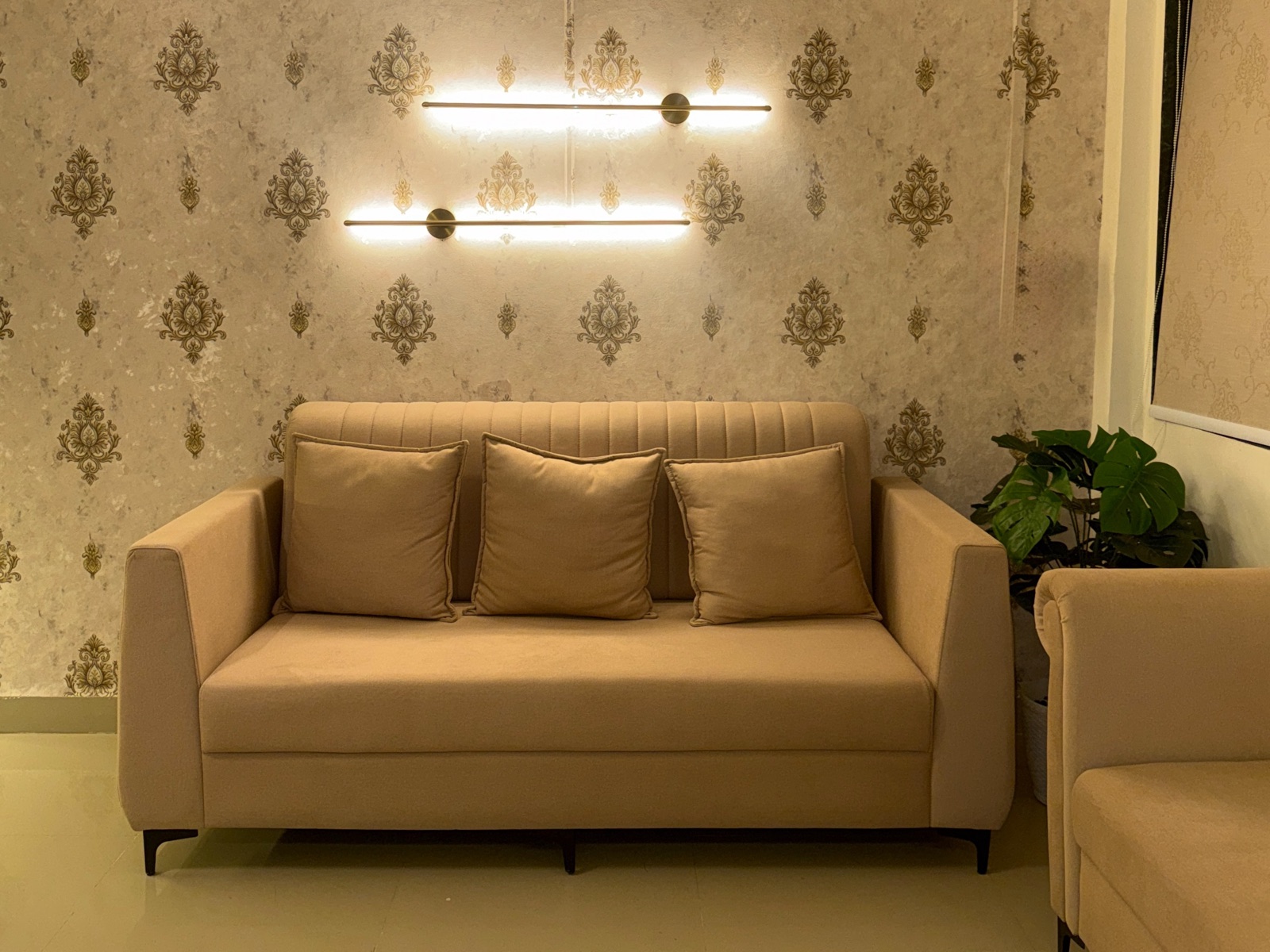 Photograph of a living room in a calm beige and cream palette: a tufted beige three-seater sofa with three cushions against a damask-patterned wallpaper wall, two linear wall lights glowing above it, a potted plant to the right and the arm of a second sofa in the foreground.
