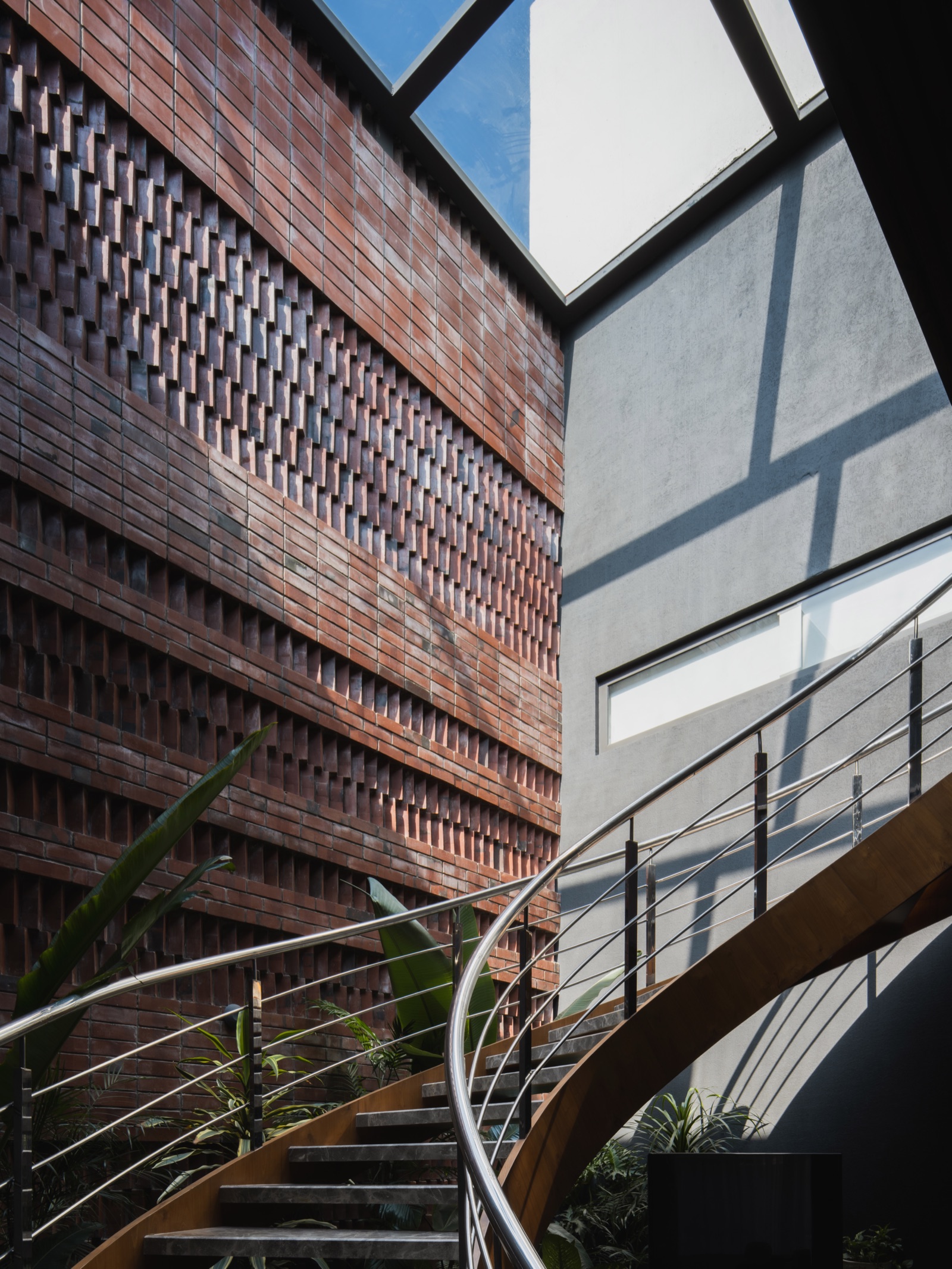Tall interior wall of red brick laid as a perforated jaali, with alternate bricks set forward and gaps between them, rising beside a curved timber and steel staircase under a large skylight, next to a smooth grey plastered wall