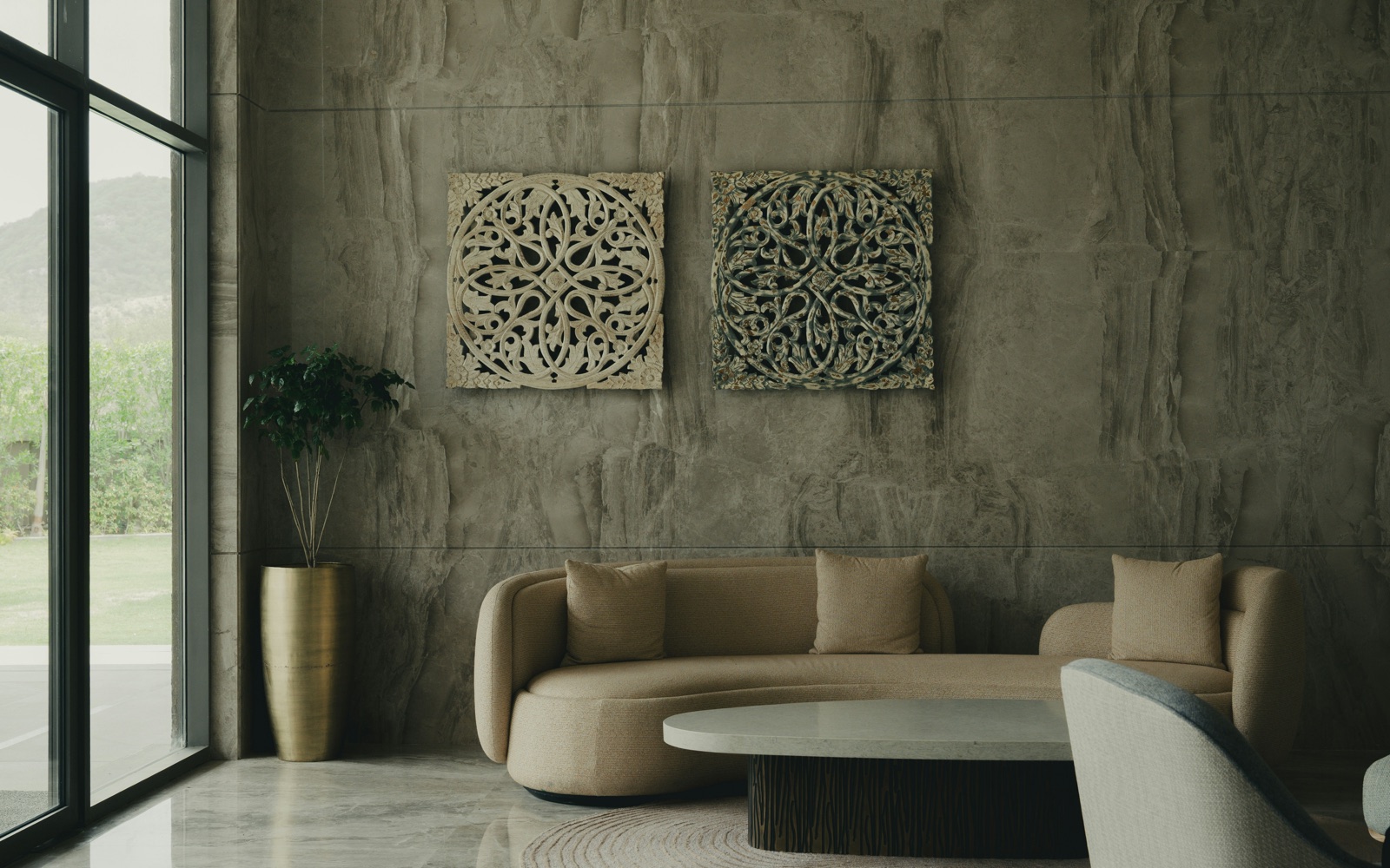 A low, curved beige sofa with textured upholstery in front of a grey stone-look wall hung with two carved wooden panels, with a brass planter, an oval stone-topped coffee table and a round rug