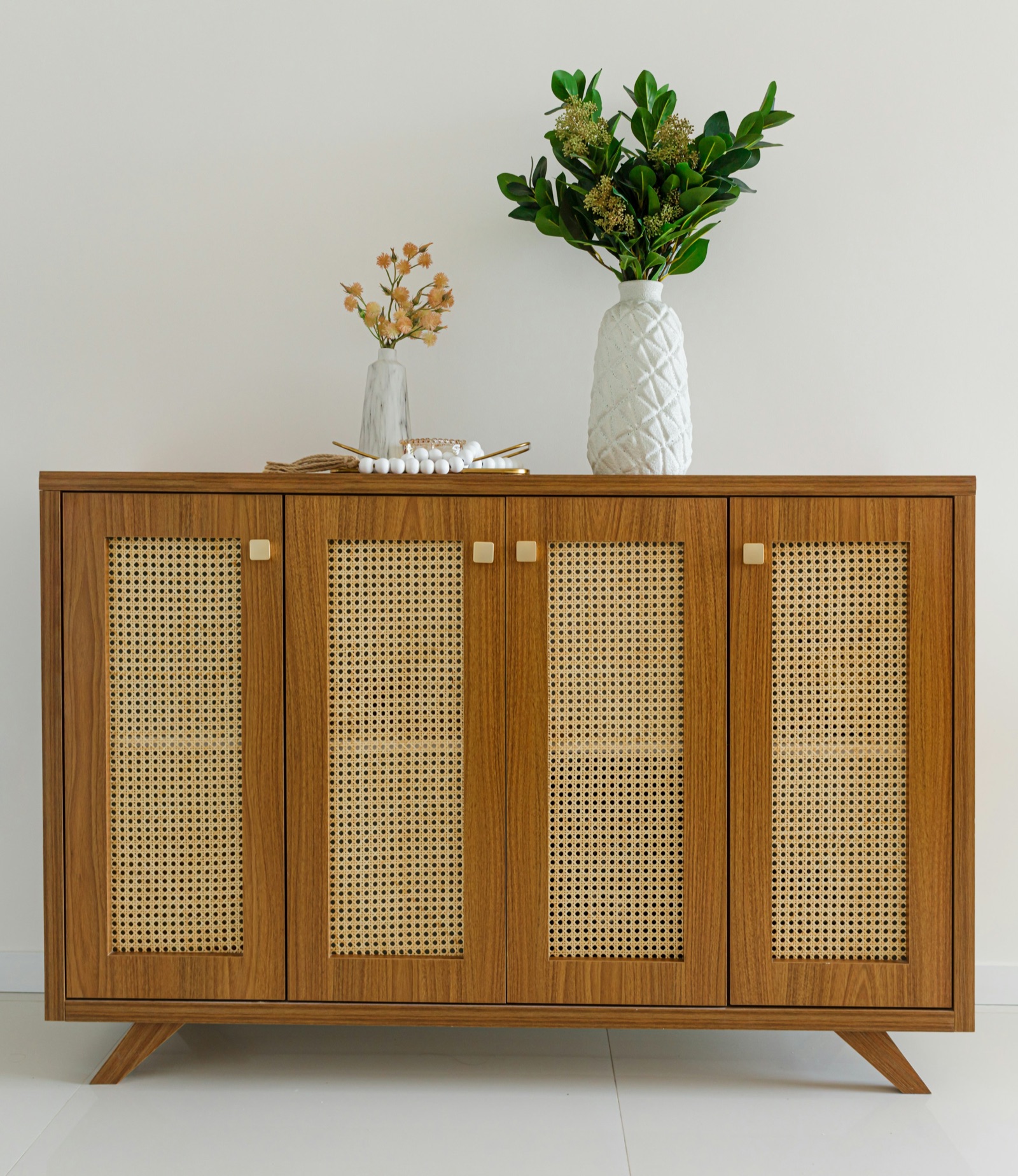 Four-door wood-finish sideboard with woven cane panels set into each door, small square brass-tone knobs and splayed legs, against a white wall with a white textured vase of green leaves and a small marble-effect vase on top