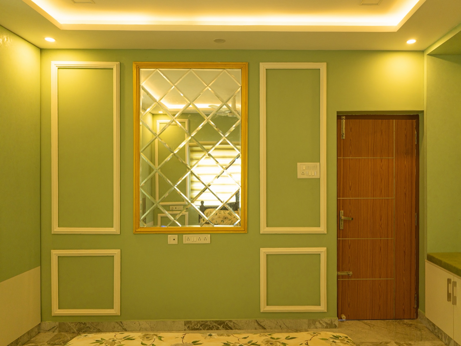 Bedroom wall in a Chennai home painted sage green, with white moulded panel frames, a gold-framed mirror of bevelled diamond-cut glass in the centre, a cove false ceiling with warm hidden lighting and downlights, and a wood-laminate door on the right