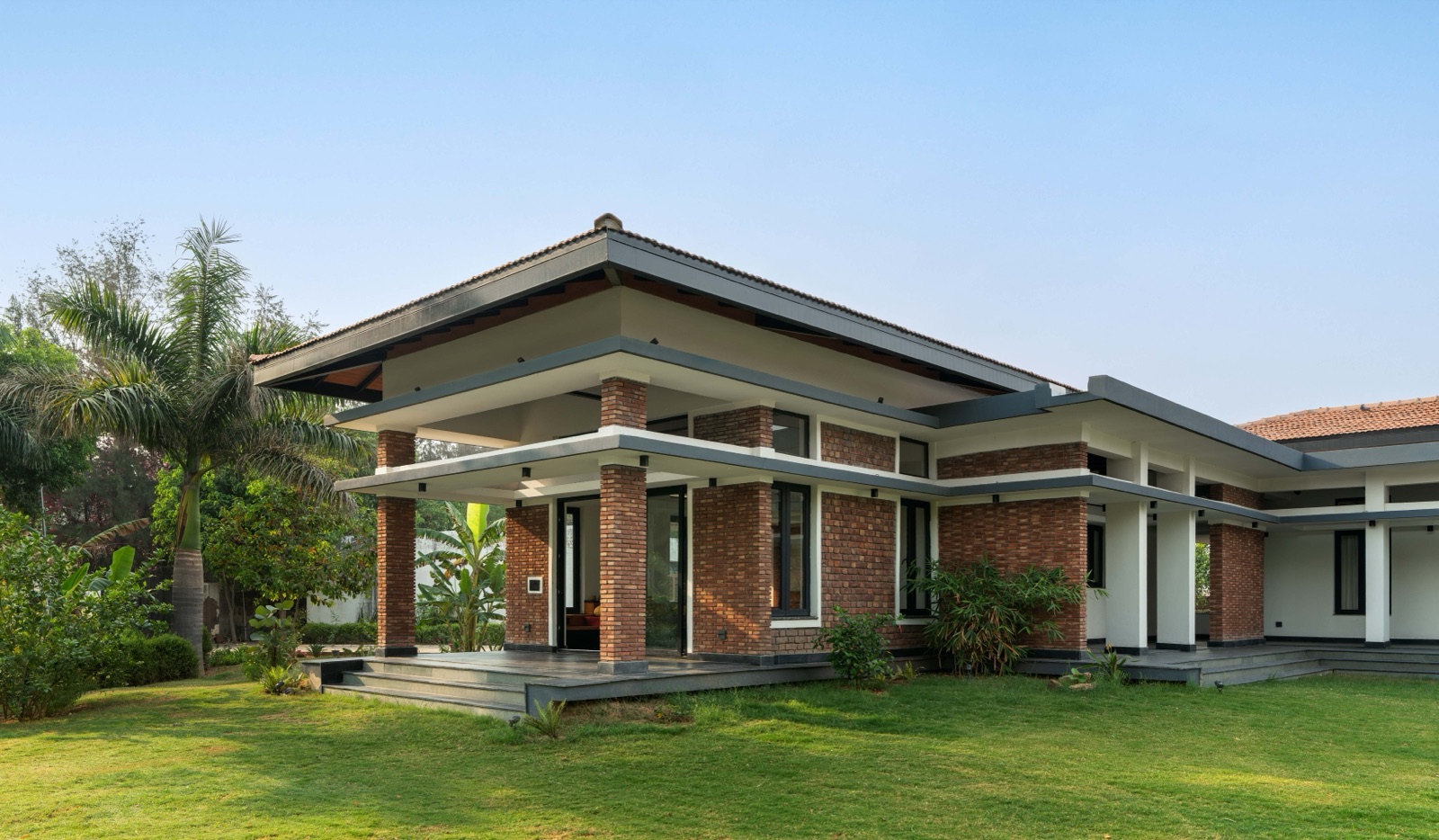 A single-storey house with exposed brick walls, slender brick piers and a broad four-way pitched roof over deep verandahs, set on a green lawn with palms on the left