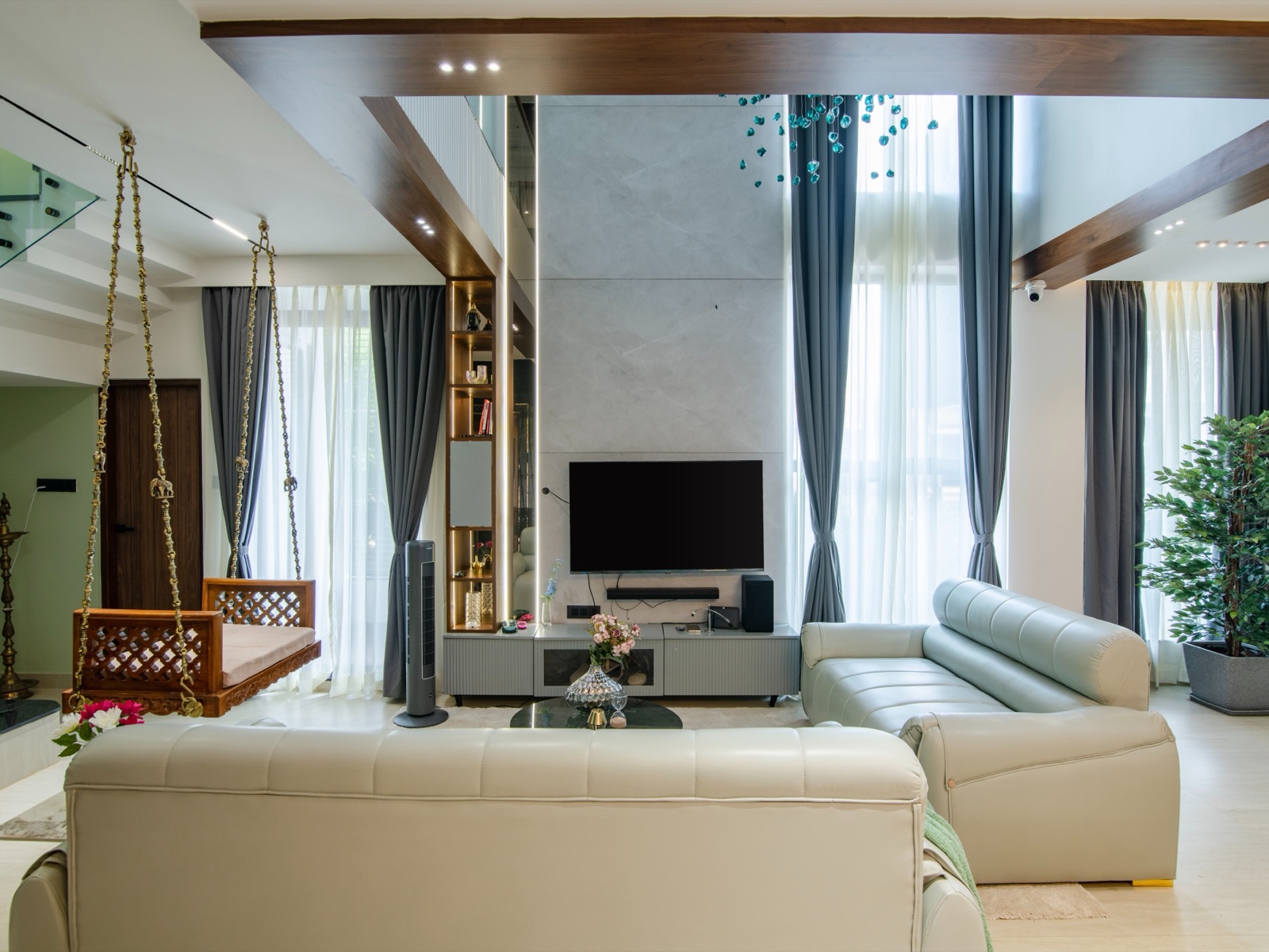 A double-height living room with a carved wooden jhoola hung on brass chains beside two cream leather sofas, a TV on a pale stone wall, full-height sheer and grey curtains and a tall brass lamp stand