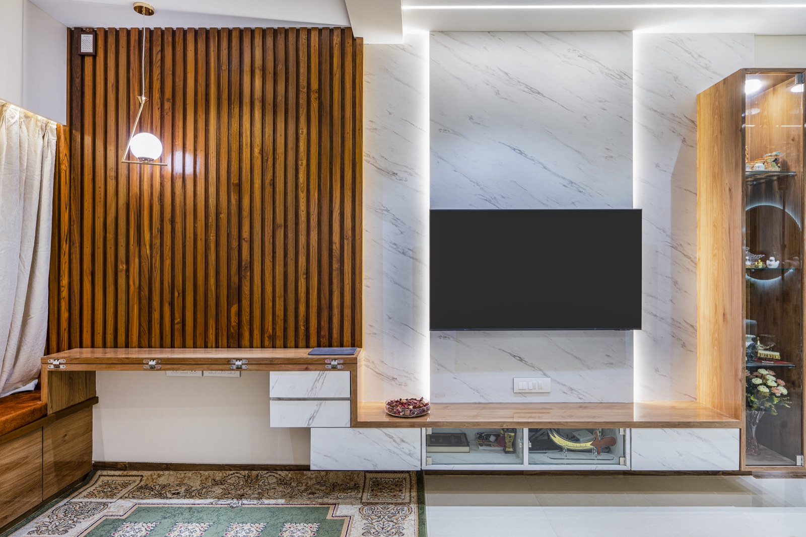 Living room wall in a Mumbai Suburban flat with vertical timber slats, a fold-down wooden desk, a backlit white marble-finish TV panel with a wall-mounted television and a tall glass display cabinet