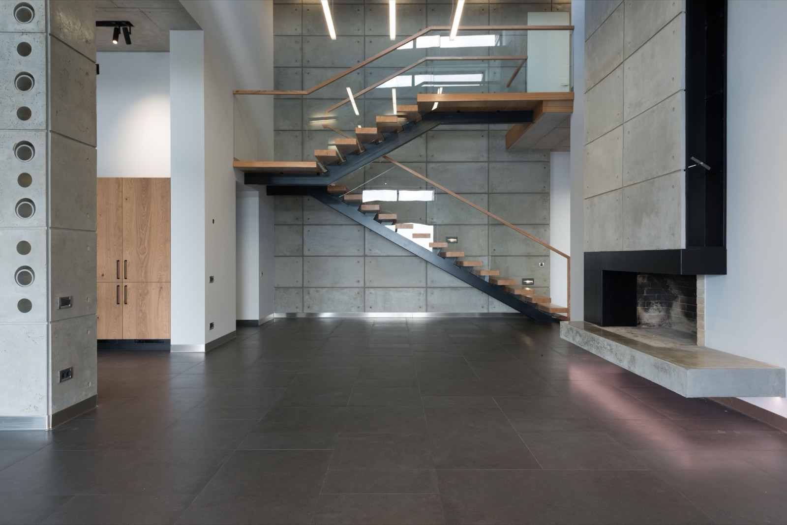 Open-plan double-height space with a concrete-panel wall and a staircase of black steel stringers carrying light oak treads, with a mid landing and a glass balustrade topped by a wooden handrail
