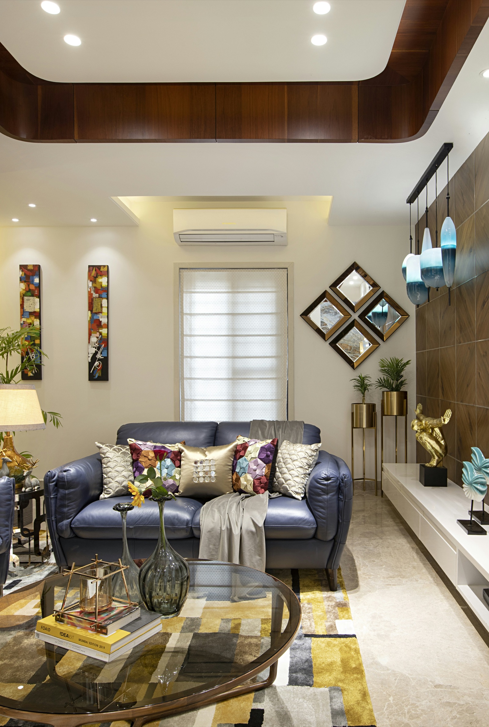 Living room with a curved false ceiling edged in dark wood veneer and recessed downlights, a blue leather sofa with patterned cushions under a window with a white roller blind, four diamond-shaped mirrors on the wall, wood-panelled wall with glass pendant lights and a pale marble-look floor