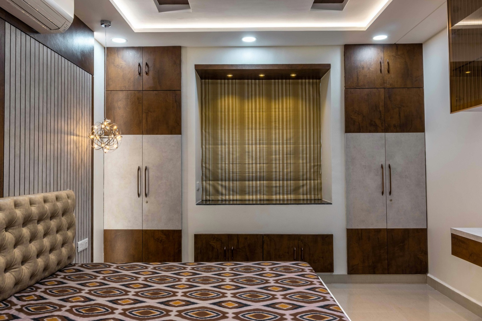 A bedroom wall with two full-height wardrobes in walnut and pale grey laminate on either side of a recessed window with a striped roman blind, a low row of drawers under the window, a split AC high on the wall to the left, a sputnik pendant light, cove lighting and downlights in the ceiling, and a tufted headboard in the foreground.