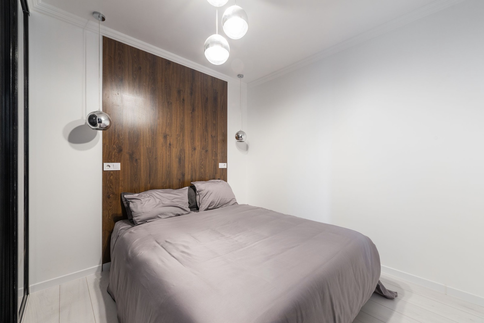 A small white bedroom with a double bed against a full-height dark wood panel, a chrome pendant light hanging low on each side of the bed instead of bedside lamps, sockets set into the panel at pillow height and the edge of a sliding wardrobe at the left