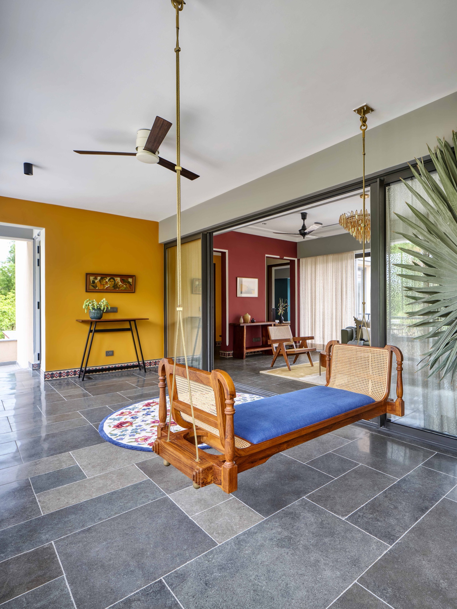 A semi-open verandah with a teak and cane swing hung from brass rods over a floor of large grey rectangular slabs laid in a random pattern with fine pale joints. A mustard wall, a console table and sliding glass doors to a living room sit behind it.
