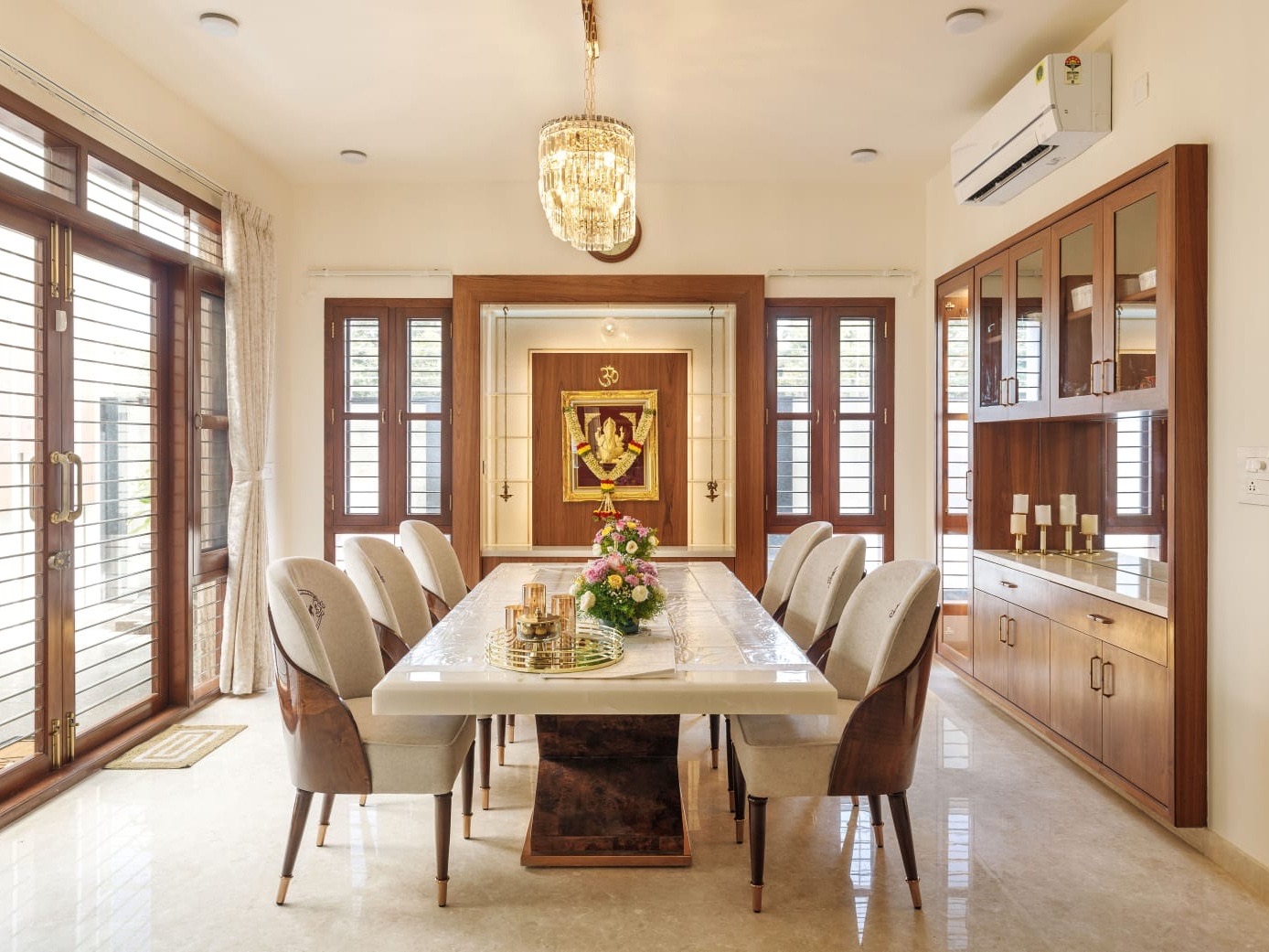 A dining area with six upholstered chairs around a white stone table, a framed Ganesha panel with a garland in a lit wooden niche between two louvred windows, a full-height wooden crockery unit with glass shutters on the right and a crystal chandelier