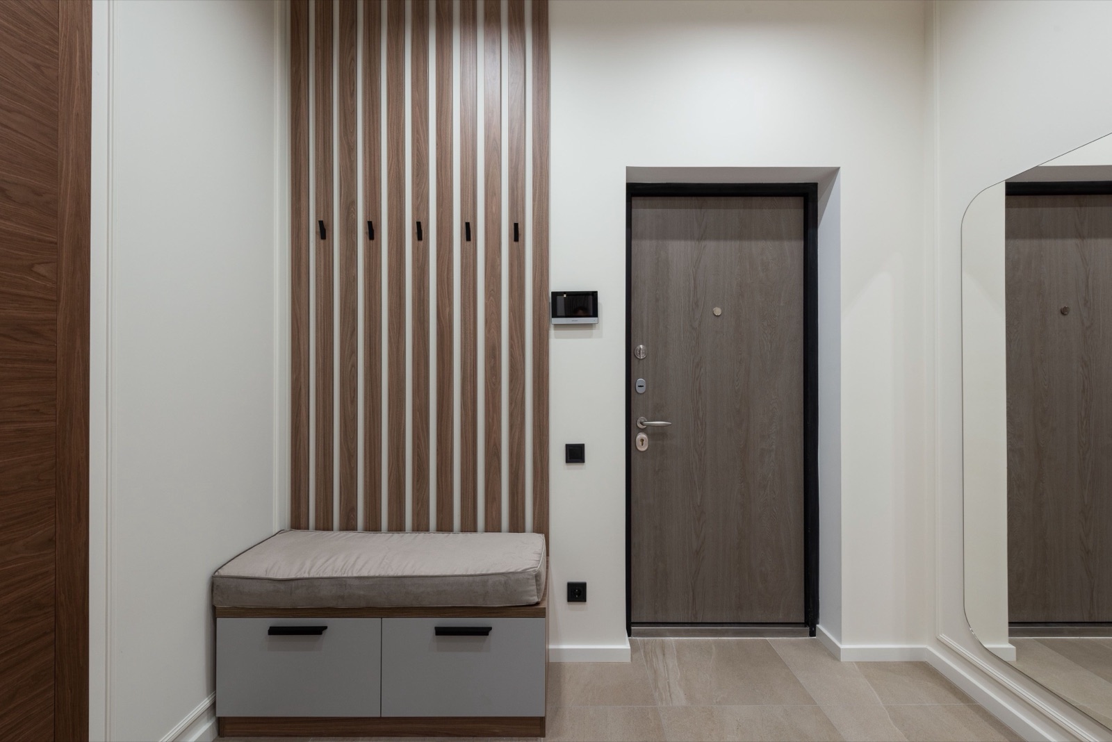 An apartment entrance with a built-in bench: a grey upholstered seat cushion on a low unit with two grey drawers, a wall of vertical wooden slats with black coat hooks above it, a wood-finish main door, a beige tiled floor and a tall mirror on the right wall