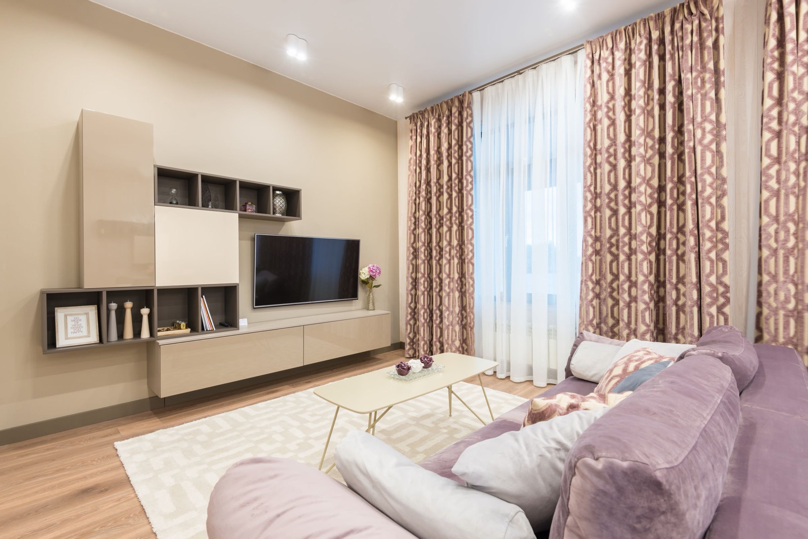Small living room with a wall-mounted TV beside a floating beige gloss storage unit made of a low drawer console, open cubbies and a tall cabinet, all lifted off the floor, with a lilac sofa and light coffee table in front