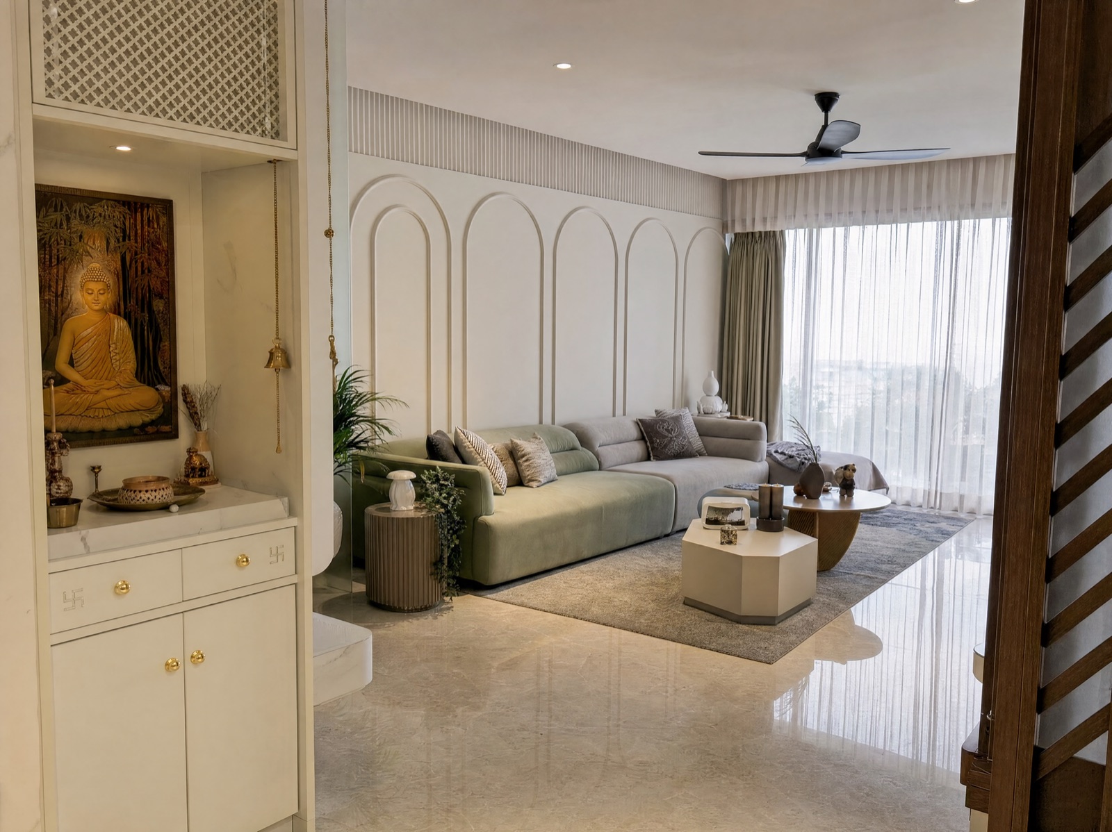 A built-in floor-standing mandir unit at the edge of a Kolkata living room: white lacquered cabinet with two drawers and two doors, small brass knobs and swastika inlays, a white marble-look top holding a brass plate, diya and small brass items, a framed painting of the Buddha on the back wall, a brass bell hanging on a chain, a recessed downlight, and a white lattice jaali panel above. Beyond it, a sage-green sofa against white arched wall panelling.
