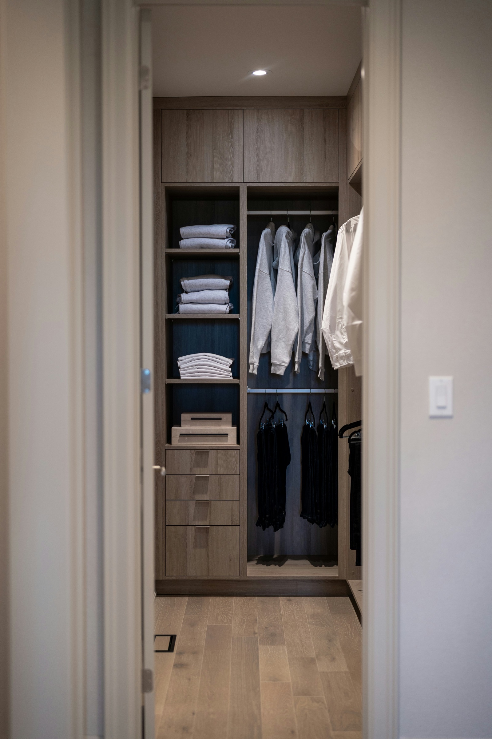 View through a doorway into a wood-finish wardrobe: one bay with open shelves of folded clothes above four drawers, and one bay with two hanging rails, light tops on the upper rail and dark garments on the lower, under closed cabinets at the top.