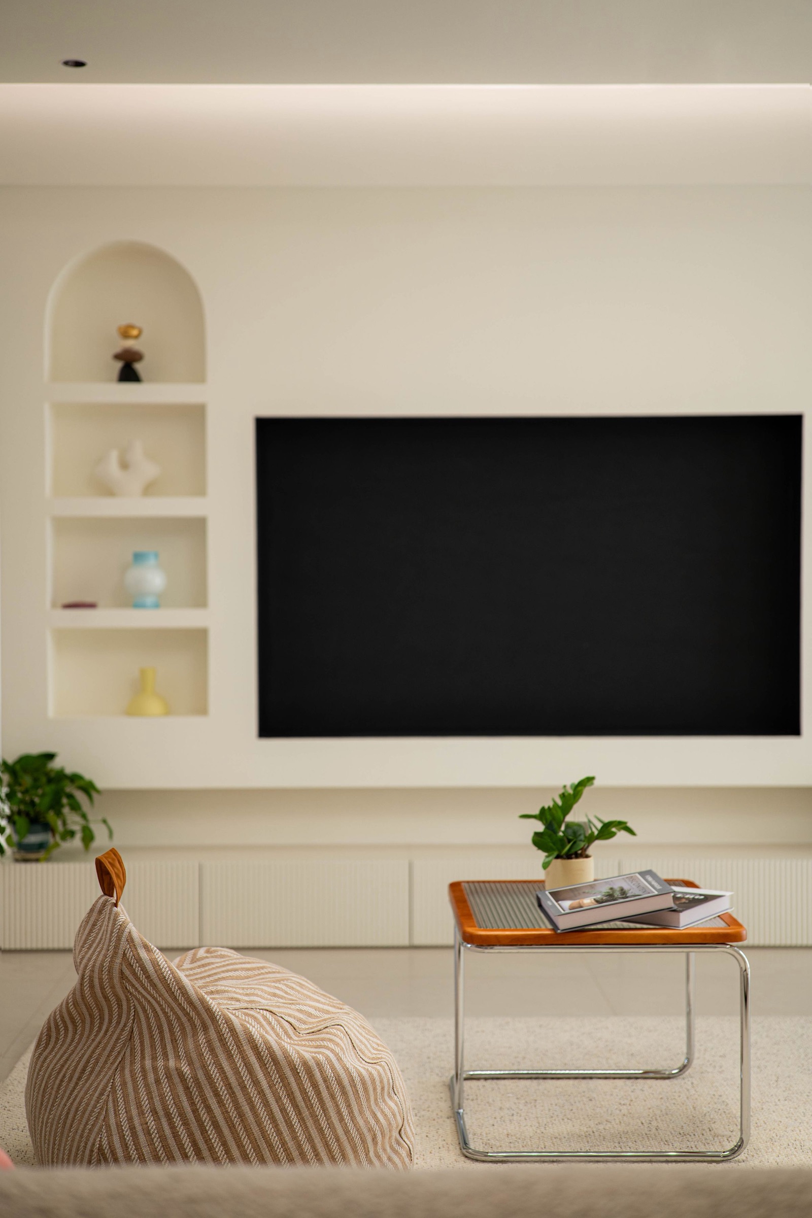 Cream living room wall with a TV set flush into a recess, a column of arched and square open niches holding vases beside it, and a long low built-in console with fluted fronts, with a striped bean bag and a small table in the foreground